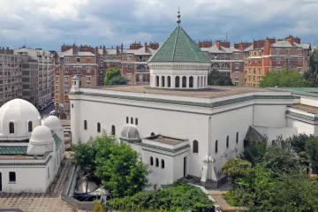L'image montre un bâtiment architectural distinctif avec une façade blanche et un toit en tuiles vertes. Le design présente des éléments inspirés de l'architecture islamique, comme des dômes et des arches. En arrière-plan, on aperçoit des immeubles de style haussmannien, typiques de nombreuses villes françaises, avec des balcons et des toits en attique. Le ciel est nuageux, ce qui ajoute une atmosphère particulière à la scène. Des arbres et de la verdure entourent le bâtiment, contribuant à l'esthétique générale.