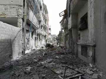 L'image montre une rue dévastée, probablement dans une zone de conflit. Les bâtiments de chaque côté sont endommagés, avec des murs effondrés et des débris éparpillés sur le sol. Des fils électriques pendent, et l'atmosphère générale dégage une impression de destruction et d'abandon. On peut observer des traces des conséquences des affrontements, avec un paysage urbain sévèrement altéré.