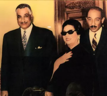 الصورة تظهر ثلاثة أشخاص يقفون معًا في إطار أنيق. الشخص الموجود على اليسار يبدو في بدلة رسمية وبابتسامة ودودة، بينما الشخصية في المنتصف ترتدي نظارات شمسية وقبعة، تبدو مميزة جدا في ملابسها، وتعبر عن شخصية قوية. الشخص الثالث على اليمين يبدو أيضًا في بدلة رسمية. الخلفية تحتوي على تصميم خشبي، مما يضيف لمسة من الفخامة إلى الصورة.
