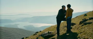 L'image montre un homme et un enfant sur une colline, face à un paysage naturel vaste et montagneux. L'homme semble parler à l'enfant, peut-être pour lui donner des conseils ou partager un moment d'intimité. En arrière-plan, on aperçoit un lac scintillant et des montagnes, le tout baigné par une lumière douce, ce qui crée une atmosphère sereine et contemplative. La scène évoque des thèmes de connexion et de découverte en pleine nature.