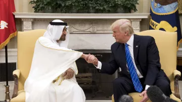 En la imagen se puede ver a dos hombres en un ambiente formal. Uno de ellos viste una túnica blanca y una kefía, que son vestimentas tradicionales de algunos países árabes, mientras que el otro lleva un traje oscuro con una camisa blanca y una corbata azul. Ambos están en un apretón de manos, lo que sugiere un acuerdo o una conversación amistosa. Al fondo, se aprecia una decoración sobria y plantas, así como banderas que indican que se encuentran en un lugar oficial, probablemente una oficina o sala de reuniones.