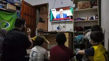 L'image montre un groupe d'enfants assis sur le sol, regardant la télévision. Un adulte, probablement un parent, est également présent et semble interagir avec les enfants. La télévision affiche un homme parlant, probablement un discours ou une intervention publique. L'environnement est simple, avec des meubles basiques et des objets éparpillés. Les enfants semblent absorbés par ce qu'ils voient à l'écran.