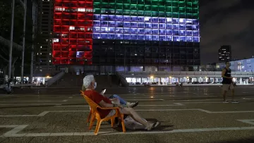 L'image montre une scène nocturne dans une ville. Au premier plan, un homme âgé est assis sur une chaise orange, en train de regarder son téléphone. En arrière-plan, un bâtiment imposant est illuminé avec des lumières de différentes couleurs, formant un motif qui ressemble à un drapeau. Les couleurs rouge, vert et bleu s'alternent sur la façade du bâtiment, créant une ambiance vibrante. D'autres personnes se déplacent dans l'espace public, ajoutant de la vie à la scène.