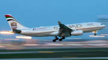 L'image montre un avion de la compagnie aérienne Etihad Airways en phase de décollage ou d'atterrissage. L'avion est partiellement flou, ce qui suggère une vitesse élevée. Le ciel apparaît dans une teinte bleue, tandis que des lumières brillantes en arrière-plan ajoutent une ambiance dynamique. L'appareil affiche la décoration et le logo caractéristiques de la compagnie.