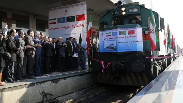 در این تصویر، یک قطار در ایستگاهی با مقامات و افراد حضور دارد. در پس‌زمینه، پرچم‌های کشورهای مختلف به چشم می‌خورد. چندین نفر در سمت چپ تصویر ایستاده و به سمت قطار توجه دارند، برخی از آنها در حال applauding هستند. تابلوهایی با متن‌هایی درباره افتتاح و همکاری‌های بین‌المللی نیز در تصویر دیده می‌شود. این صحنه نشان‌دهنده یک رویداد ویژه یا افتتاحیه مرتبط با حمل و نقل ریلی است.