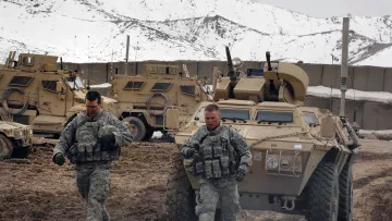 تظهر الصورة مشهدًا عسكريًا في منطقة جبلية. في الصورة، يسير جنديان في زي عسكري في المقدمة، مع التركيز على تفاصيل ملابسهم ومعداتهم. خلفهما، توجد مركبات مدرعة تُستخدم في العمليات العسكرية، محاطة بتضاريس جبلية تغطيها الثلوج. يبدو أن الطقس بارد، ويعكس المشهد البيئة الصعبة التي يعمل فيها الجنود.