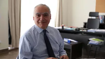 L'immagine mostra un uomo sorridente seduto a un tavolo in un ambiente di lavoro. Indossa una camicia e una cravatta, e sembra essere concentrato su alcune carte e documenti posti davanti a lui. Sullo sfondo, si intravedono elementi di un ufficio, come una scrivania con vari materiali e un ambiente luminoso grazie alla luce naturale che entra dalla finestra.