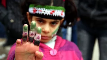 L'image montre une jeune fille qui lève la main en avant, exhibant ses doigts teintés de peinture. Elle porte un pull en laine rose et un bandeau coloré orné de motifs. Son visage est marqué par des traces de peinture, notamment sur les joues. L'expression de son visage semble déterminée, et elle attire l'attention sur son geste. En arrière-plan, on peut apercevoir des silhouettes floues de personnes, suggérant un rassemblement ou une manifestation.