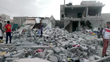 L'image montre des bâtiments gravement endommagés, avec des débris éparpillés sur le sol. On peut voir des enfants et des adultes présents parmi les ruines, cherchant peut-être des effets personnels ou évaluant les dégâts. L'environnement semble chaotique et témoigne d'une destruction significative, probablement suite à un conflit ou à une catastrophe. Le ciel est nuageux, ce qui ajoute à l'atmosphère sombre de la scène.