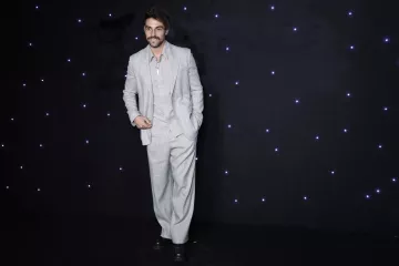 L'immagine mostra un uomo in piedi su uno sfondo scuro, illuminato da piccole luci bianche che brillano. Indossa un completo grigio chiaro, caratterizzato da pantaloni ampi e una giacca ben tagliata. La sua camicia è di un tono chiaro e ha il colletto aperto. L'uomo ha un sorriso e porta dei baffi, dando un'aria elegante e sicura di sé.