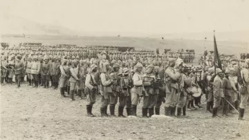 L'image montre une grande formation de soldats en uniforme, probablement d'un conflit militaire historique. Ils sont alignés en rangées serrées sur un terrain désertique ou peu végétalisé. Certains soldats portent des armes et semblent être en position de marche ou de formation. En arrière-plan, on peut voir d'autres soldats formant également des rangées, ce qui donne une impression de mobilisation de grandes troupes. L'atmosphère est sérieuse et solennelle, évoquant le contexte militaire de l'époque.
