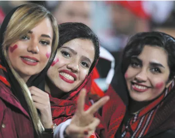 L'image montre trois femmes souriantes, vêtues de vêtements dans des tons sombres, avec des écharpes ou des foulards sur la tête. Chacune d'elles a des cœurs rouges dessinés sur les joues, ce qui suggère qu'elles célébrent un événement sportif ou une occasion festive. Elles affichent des expressions de joie et de camaraderie, et l'une des femmes montre un signe de paix avec ses doigts. L'arrière-plan est flou, mais on peut discerner une ambiance festive.