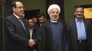 L'image montre trois hommes se tenant debout, avec un homme au centre qui sourit et porte une robe traditionnelle, tandis que les deux autres sont en costume. Ils semblent être en train de se saluer, probablement lors d'une rencontre officielle. L'environnement semble formel, probablement dans un hôtel ou un bâtiment gouvernemental, avec des personnes en arrière-plan.