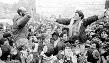 L'image montre une foule de personnes rassemblées, avec des visages enthousiastes et des expressions de joie. Deux hommes se distinguent, l'un portant des lunettes, debout sur les épaules de l'autre, qui lève les bras en signe de célébration. Les membres de la foule semblent excités et engagés, et l'environnement est peut-être urbain ou rural, entouré de bâtiments en pierre. L'atmosphère générale évoque un moment de rassemblement collectif et de camaraderie.