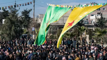 L'image montre une foule rassemblée sous de grandes drapeaux verts et jaunes, portant une étoile rouge. En arrière-plan, des bâtiments et des arbres sont visibles. Des banderoles sont accrochées au-dessus des personnes, indiquant une célébration ou un rassemblement communautaire. L'atmosphère semble vibrante et engagée, avec un grand nombre de participants.