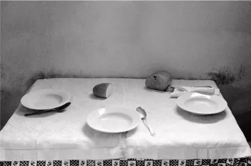 L'image montre une table couverte d'une nappe blanche, où se trouvent deux assiettes blanches, une cuillère et un couteau. Un petit pain est positionné sur un côté et un verre est renversé au centre de la table. L'ambiance générale est sobre et monochrome, ce qui donne une impression de simplicité et de calme. Les murs derrière la table sont légèrement ombragés, ajoutant un effet de profondeur à la composition.