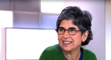 L'image montre une femme souriante avec des cheveux gris et courts. Elle porte des lunettes et un pull de couleur verte. Son expression est amicale et chaleureuse, indiquant qu'elle participe à une conversation. L'arrière-plan est flou, suggérant qu'elle se trouve dans un environnement de studio ou d'interview.