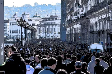 L'image montre une foule dense de personnes marchant dans une rue, probablement dans une ville animée. Les bâtiments sur les côtés semblent historiques, avec une architecture riche. On peut voir des lampadaires le long de la promenade, et au loin, on aperçoit des collines ou des montagnes qui ajoutent à la profondeur de la scène. L'atmosphère semble vibrante et dynamique, reflétant probablement une manifestation ou un événement public.