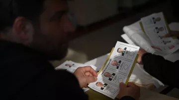 En la imagen se puede ver a una persona sosteniendo una boleta electoral. La boleta muestra varias fotografías de candidatos, junto con sus nombres y posiblemente posiciones a las que aspiran. La persona parece estar concentrada en la boleta, mientras que en la mesa hay más documentos. La escena parece tener lugar en un ambiente de votación.