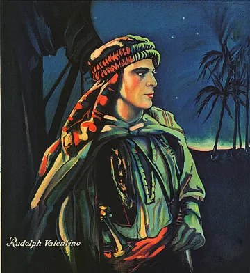 الصورة تظهر شخصية ترتدي زيًا تقليديًا، مع غطاء رأس مزخرف. تبدو الشخصية جادة وتحدق بعيدًا، بينما خلفها سماء مظلمة مملوءة بالنجوم. هناك أشجار من نوع النخيل تظهر في الخلفية، مما يضيف إلى الجو الغامض للصورة. الاسم "Rudolph Valentino" مكتوب في الزاوية، وهو يشير إلى شخصية معروفة في عالم السينما. الألوان المستخدمة في الصورة تعكس تدرجات دافئة، مما يضيف عمقاً للتعبير.