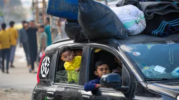 L'immagine mostra un'auto con un carico sopra di sacchi neri e altri oggetti. All'interno della vettura, si vedono due bambini: uno che guarda fuori dal finestrino con espressione curiosa e l'altro che si affaccia dall'apertura. Sullo sfondo si possono notare altre persone e un ambiente urbano, che sembra un'area affollata. La scena riflette una situazione di vita quotidiana in un contesto urbano.