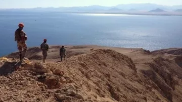 L'image montre un paysage désertique avec des militaires en patrouille sur une crête surplombant une étendue d'eau. Le terrain est aride et rocheux, avec des montagnes en arrière-plan. Les soldats portent des uniformes et des casques, et semblent observer l'horizon, avec un ciel clair et ensoleillé qui ajoute à l'atmosphère de la scène.