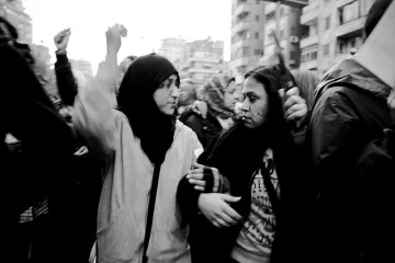 L'image montre un groupe de personnes lors d'une manifestation. Deux jeunes femmes, l'une portant un hijab et l'autre les cheveux détachés, semblent échanger des mots tout en levant les bras, ce qui indique un sentiment d'engagement et de solidarité. L'ambiance est dynamique, et le noir et blanc donne une atmosphère intense à la scène. En arrière-plan, on peut apercevoir d'autres manifestants, créant une impression de mouvement et de rassemblement.