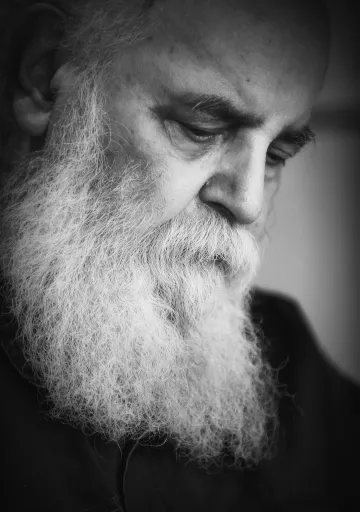 L'image montre un homme âgé avec une longue barbe blanche. Son expression est pensive et introspective, les yeux baissés. L'éclairage est doux, créant un contraste qui met en valeur les détails de son visage et de sa barbe. L'ensemble donne une ambiance de réflexion et de sérénité. La photo est en noir et blanc, ce qui accentue la gravité et la profondeur des émotions.