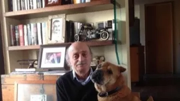 L'image montre un homme assis sur le sol, avec un chien à ses côtés. L'homme porte un pull noir et a des cheveux grisonnants. Le chien, de taille moyenne, a un pelage brun et semble calme. En arrière-plan, on peut voir une bibliothèque remplie de livres, ainsi que quelques objets personnels comme des cadres photo et des souvenirs. L'ambiance de la pièce paraît chaleureuse et accueillante.