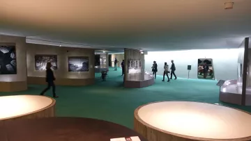 The image depicts an interior space, likely an art gallery or museum. The floor is covered with green carpeting, and the walls display a series of black-and-white photographs. Several people are walking through the space, engaged with the exhibits. There are curved display structures that showcase the photographs. The lighting appears bright and modern, creating an inviting atmosphere.