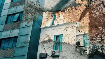 La imagen muestra la fachada de un edificio moderno de varios pisos, con ventanas de cristal y estructura metálica. Al lado, hay una pared de una construcción más antigua, que presenta desconchones y una textura envejecida. Se pueden ver persianas de color azul en algunas ventanas y un aire acondicionado montado en la pared. La mezcla de elementos arquitectónicos modernos y antiguos crea un contraste visual interesante, resaltando la historia y evolución del entorno urbano.