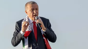 L'image montre un homme en train de parler au micro, probablement lors d'un discours ou d'une manifestation publique. Il porte un costume sombre avec une cravate rouge et un foulard aux motifs représentant la Palestine, qui est enroulé autour de son cou. L'arrière-plan semble clair, suggérant qu'il se trouve à l'extérieur. L'homme semble exprimer des émotions fortes, ce qui indique qu'il est engagé dans son discours.