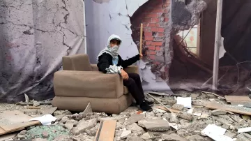 L'image montre une scène de destruction avec des débris éparpillés au sol, tels que des morceaux de béton et des morceaux de verre brisé. Au centre, une personne est assise sur un canapé apparemment plus ou moins intact. Elle porte un foulard autour de la tête et tient un bâton. En arrière-plan, il y a une illustration d'un mur brisé, ajoutant à l'impression de désolation et de chaos de la scène.