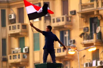 L'image montre un homme brandissant un drapeau noir, blanc et rouge, probablement un drapeau national. Il se tient debout sur une structure, manifestant une posture de célébration ou de protestation. En arrière-plan, on peut apercevoir des bâtiments avec des balcons et des climatiseurs, ce qui suggère un environnement urbain. L'atmosphère semble chargée d'une forte émotion, possiblement liée à un événement politique ou social.