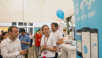 L'image montre une scène animée dans un événement, probablement un salon ou une foire. On peut voir des personnes interagissant autour d'un stand. Un homme, habillé en chemise blanche, semble discuter avec une jeune femme qui porte un uniforme avec un logo. À l'arrière-plan, il y a une bannière portant le logo d'Engie et des représentations d'applications sur des smartphones. L'ambiance est conviviale, avec des gens qui semblent engagés et intéressés par ce qui se passe.