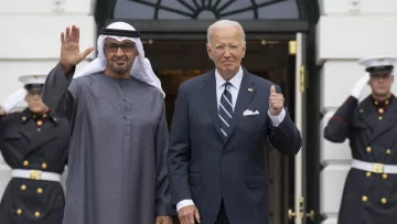 The image shows two men standing at the entrance of a building, likely the White House. One man is dressed in traditional attire with a white headscarf, while the other is in a formal suit. They are both smiling and posing for the camera, with one man waving and the other giving a thumbs-up. In the background, there are uniformed guards standing at attention. The scene conveys a sense of diplomacy and cordiality between the two leaders.