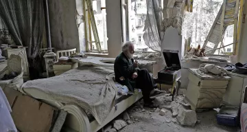 L'image montre un espace intérieur dévasté, probablement un appartement. Au centre, un homme âgé est assis sur un lit, tenant un objet à la main, peut-être une cigarette. L'environnement est marqué par des murs endommagés, des débris éparpillés et des fenêtres brisées, laissant voir l'extérieur. La lumière naturelle pénètre par les ouvertures, révélant l'état de désolation de la pièce. L'ambiance est sombre et mélancolique, illustrant les conséquences d'un conflit ou d'une catastrophe.