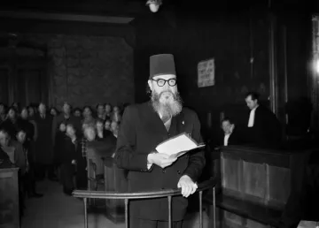 L'image montre un homme portant un turban noir et des lunettes, se tenant debout dans une salle d'audience. Il lit un document, tandis qu'un groupe de personnes l'écoute attentivement en arrière-plan. L'environnement est sombre, avec des boiseries et des bancs de juge visibles. Une personne en toge est également présente, ajoutant à l'atmosphère judiciaire de la scène.