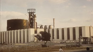 The image depicts an industrial facility, featuring a large, cylindrical structure that resembles a tank, alongside several tall chimneys and towers. The building has a series of white, vertical panels, giving it a modern appearance. In the foreground, a person on a bicycle is riding past the facility, while there are some vehicles and a tree visible. The sky has a few clouds, suggesting it might be a clear day. Overall, the scene conveys an industrial landscape with a blend of architecture and manufacture.