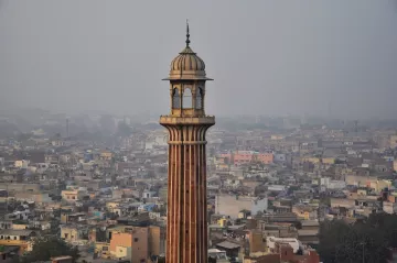 L'image montre un paysage urbain avec une tour élancée qui se dresse au milieu de nombreux bâtiments. La tour a une architecture distinctive, avec des motifs ornés et un dôme en haut. En arrière-plan, on peut voir une vaste étendue de maisons et de structures, témoignant d'une villeDense. L'atmosphère semble légèrement brumeuse, suggérant une pollution ou une humidité ambiante. Les couleurs des bâtiments varient, ajoutant une touche de diversité à l'ensemble de la scène.