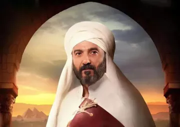 L'image représente un homme portant un turban et une tenue traditionnelle. Il a une barbe et une expression sérieuse. En arrière-plan, on aperçoit un paysage désertique avec des pyramides, suggérant une ambiance historique ou culturelle. La lumière semble douce, ajoutant une atmosphère dramatique à la scène.
