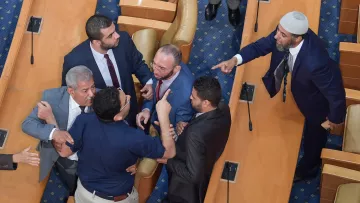 The image shows a heated confrontation occurring in a legislative assembly. Several men are engaged in a tense discussion or argument, with some gesturing animatedly. The setting appears to be a formal chamber, indicated by the rows of seats and the official appearance of the individuals. Some men are standing closely together, while others seem to be pointing or raising their hands, indicating strong emotions. Overall, the scene conveys a moment of conflict or debate among the legislators.