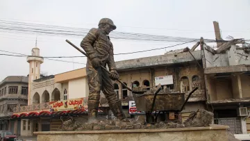 L'image montre une statue monumentale représentant un travailleur en train de creuser, symbolisant probablement la reconstruction ou le labeur dans un contexte difficile. En arrière-plan, on distingue des bâtiments partiellement détruits et des infrastructures urbaines. La scène évoque une atmosphère de résilience et de reconstruction dans une zone touchée par des conflits. La statue est installée sur un socle en pierre, ajoutant à son importance et à son impact visuel.