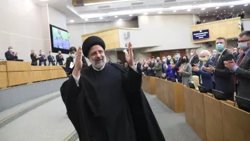 L'image montre un homme en habit traditionnel, portant un turban noir, qui semble être en train de saluer un public. Il se tient au centre d'une salle où plusieurs personnes applaudissent. L'environnement est formel, typique d'un événement politique ou d'une réunion officielle, avec des sièges et des écrans visibles en arrière-plan. Les participants portent également des masques de protection, indiquant une attention à la santé publique.