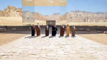 L'immagine mostra un gruppo di uomini vestiti con abiti tradizionali, dimostrando un'atmosfera di formalità e celebrazione. Sono disposti in fila su una pavimentazione di pietre chiare, con un paesaggio desertico sullo sfondo. Dietro di loro si erge una struttura riflettente che sembra integrarsi con l'ambiente circondante, creando un gioco di luci e riflessi. Le montagne in lontananza aggiungono profondità alla scena, conferendo un senso di grandiosità e atmosfera unica.