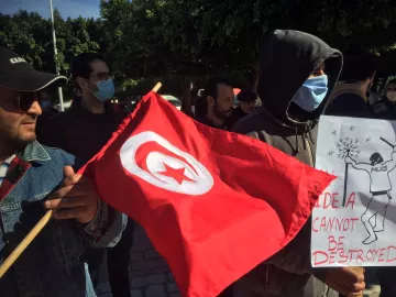 En la imagen se puede ver a un grupo de personas participando en una manifestación. Uno de los manifestantes sostiene una bandera roja con una estrella y una media luna blancas, símbolo nacional de Túnez. Otro individuo lleva un cartel que dice "Una idea no puede ser destruida", acompañado de una ilustración. La mayoría de los participantes utilizan mascarillas, y el ambiente parece ser de protesta pacífica. El fondo muestra árboles y un entorno urbano.