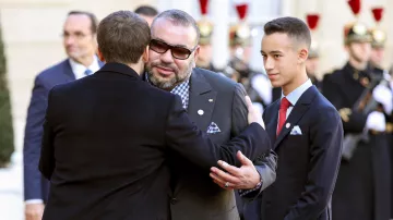 L'image montre une rencontre officielle entre deux hommes. L'un des hommes, vêtu d'un costume élégant, semble accueillir l'autre avec une accolade amicale. L'autre homme, portant des lunettes de soleil et un costume gris, se tient à côté d'un jeune garçon en costume sombre et cravate. En arrière-plan, on aperçoit des gardes en uniforme, ce qui indique un contexte cérémonial. L'atmosphère semble conviviale et respectueuse.