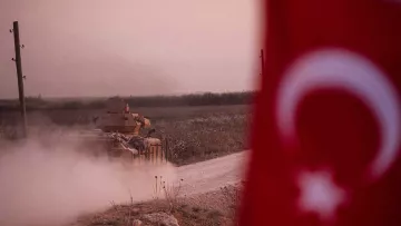 در این تصویر، یک تانک نظامی در حال حرکت در یک جاده خاکی به سمت جلو دیده می‌شود. در پس‌زمینه، میدان‌های سبز و طبیعتی آرام قرار دارد. در سمت چپ تصویر، پرچم ترکیه با نشانه‌های مشخص آن قرار دارد. غبار و دود ناشی از حرکت تانک فضای تصویر را پوشانده و حس تحرک و فعالیت نظامی را منتقل می‌کند. نور ملایم غروب یا صبح نیز به فضای کل تصویر جذابیت بیشتری بخشیده است.
