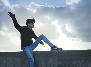 L'image montre un jeune homme assis sur un muret, avec une posture décontractée. Il porte un sweat noir et un jean bleu, avec des baskets noires. Ses cheveux sont légèrement ébouriffés par le vent, et il semble apprécier le moment en regardant vers le ciel, qui est nuageux avec quelques éclaircies. L'ambiance générale évoque une sensation de liberté et de fraîcheur.