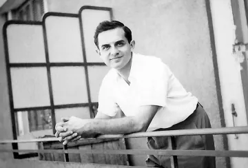 The image features a young man with a pleasant expression, leaning on a railing. He has dark hair styled in a neat manner and is dressed in a light-colored short-sleeve shirt paired with trousers. The background suggests a balcony or outdoor setting, with a partially visible modern-style partition or screen. The photo has a vintage quality, indicating it was likely taken several decades ago.